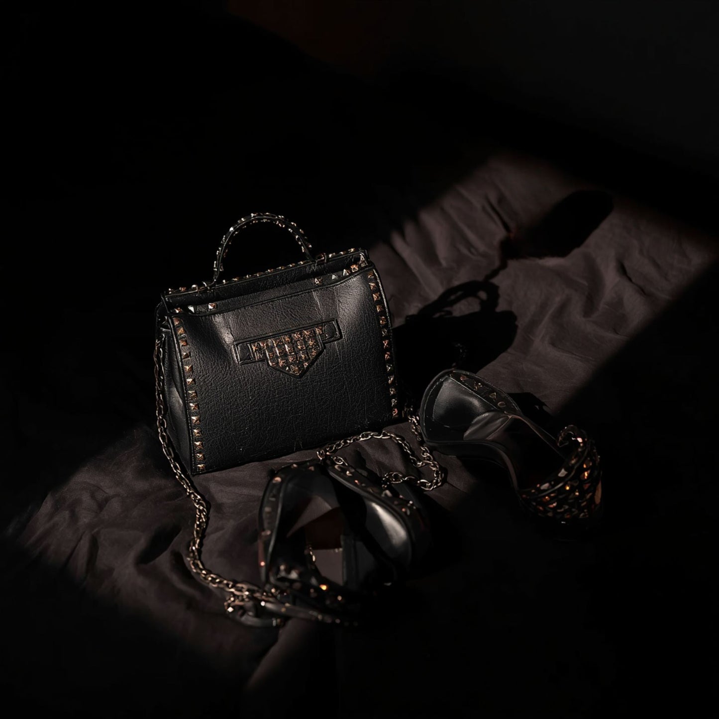 A black studded handbag and black high‑heeled shoes arranged on a dark fabric surface, lit by warm dramatic lighting that highlights the metallic studs and leather textures.