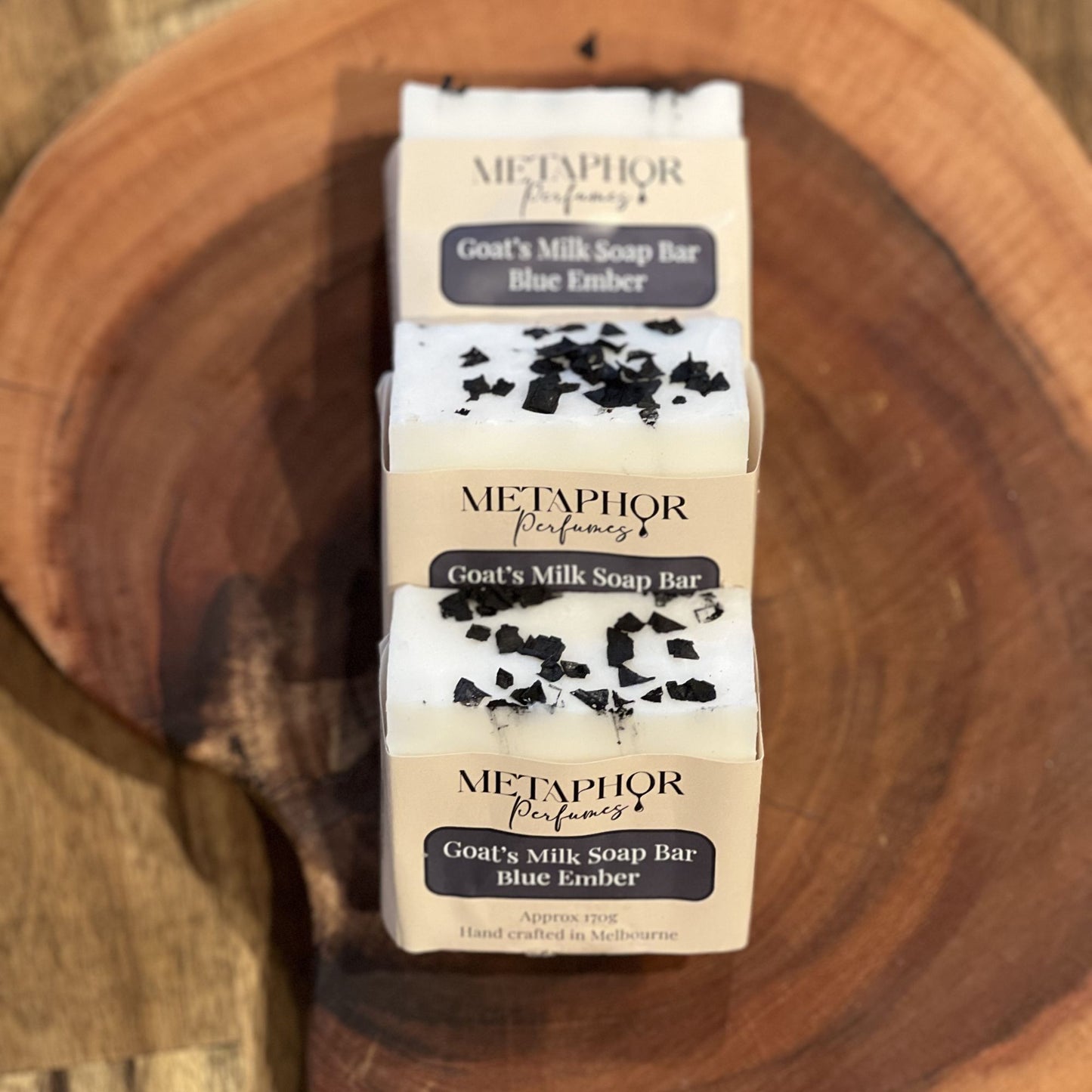 White goat’s milk soap bars with black salt flakes tops wrapped in Metaphor Perfumes Blue Ember labels, stacked on a wooden surface; inspired by Dior Sauvage