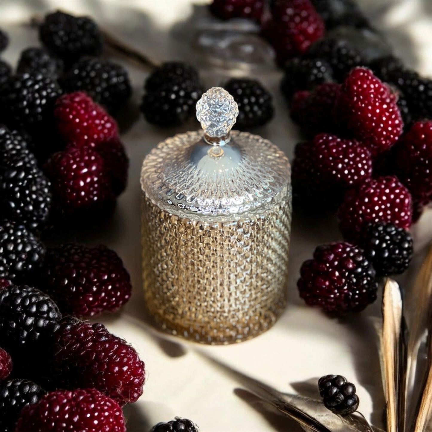 A textured golden glass candle jar with a clear faceted lid, surrounded by fresh blackberries and raspberries, representing the Black Raspberry & Vanilla scent.
