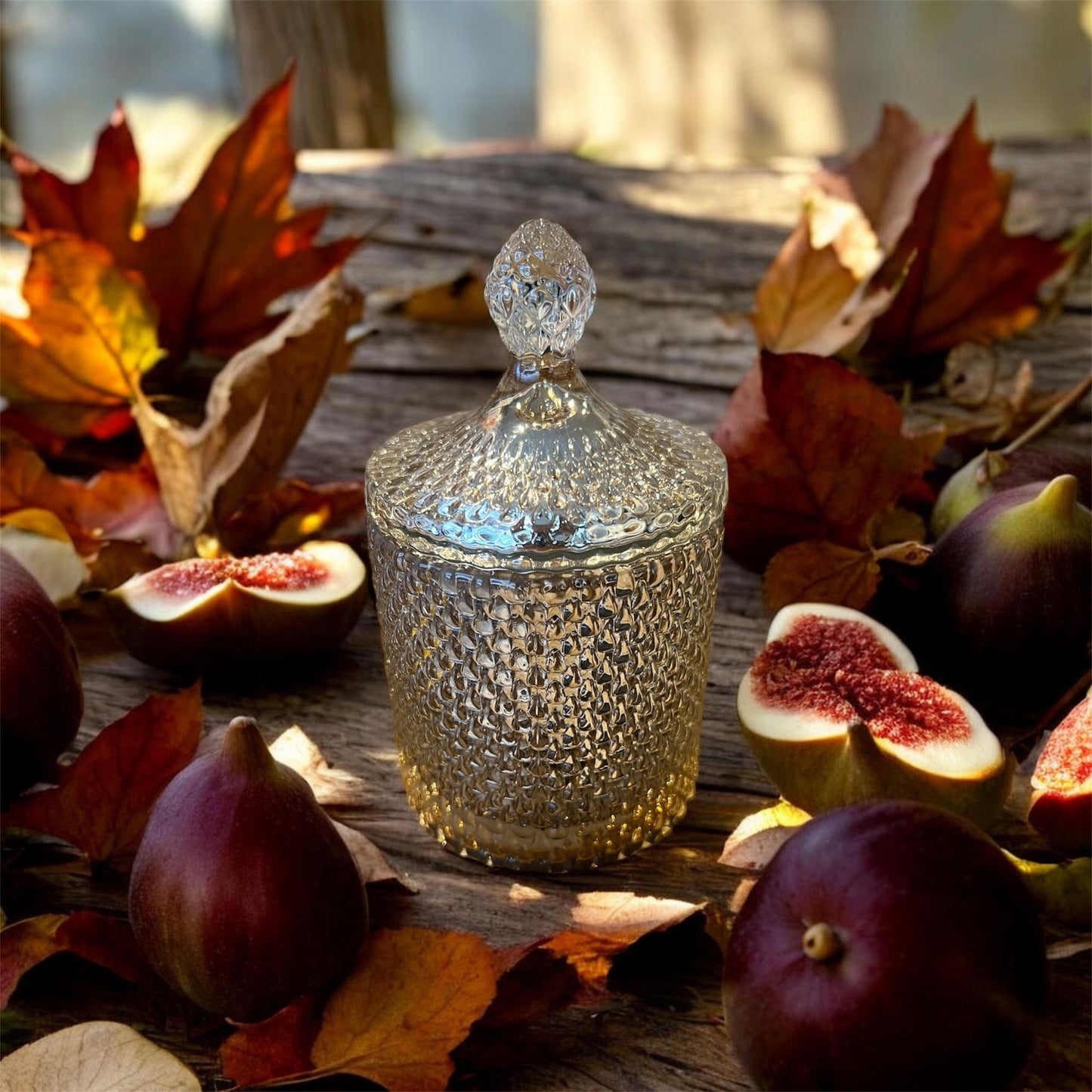 A textured glass candle jar with a faceted lid sits on a wooden surface surrounded by whole and sliced figs and scattered autumn leaves.
