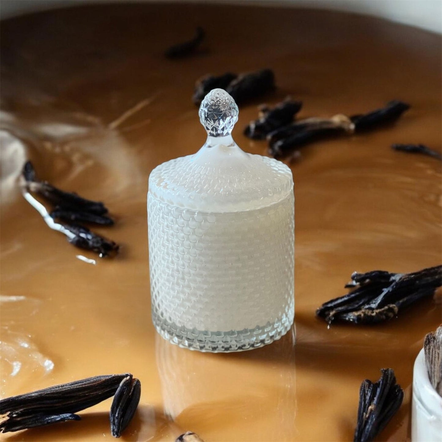 A textured glass candle jar resting on a glossy caramel surface, surrounded by dried vanilla beans, representing the Caramel Vanille candle.