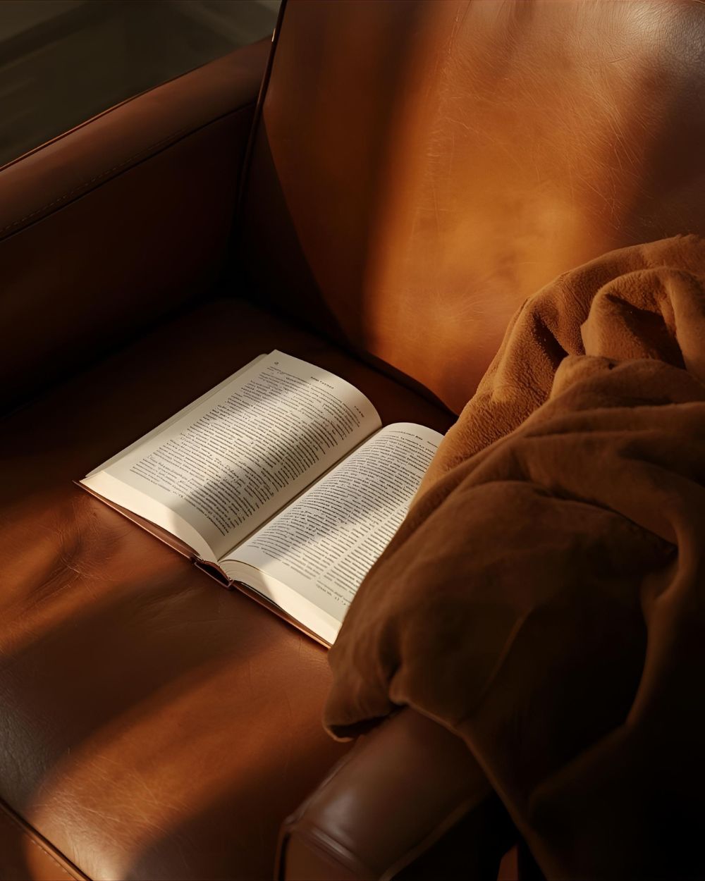 Leather extrait collection — warm, textured scents with suede, rose, musk, and spice. A brown leather armchair with an open book and draped fabric in soft morning light.