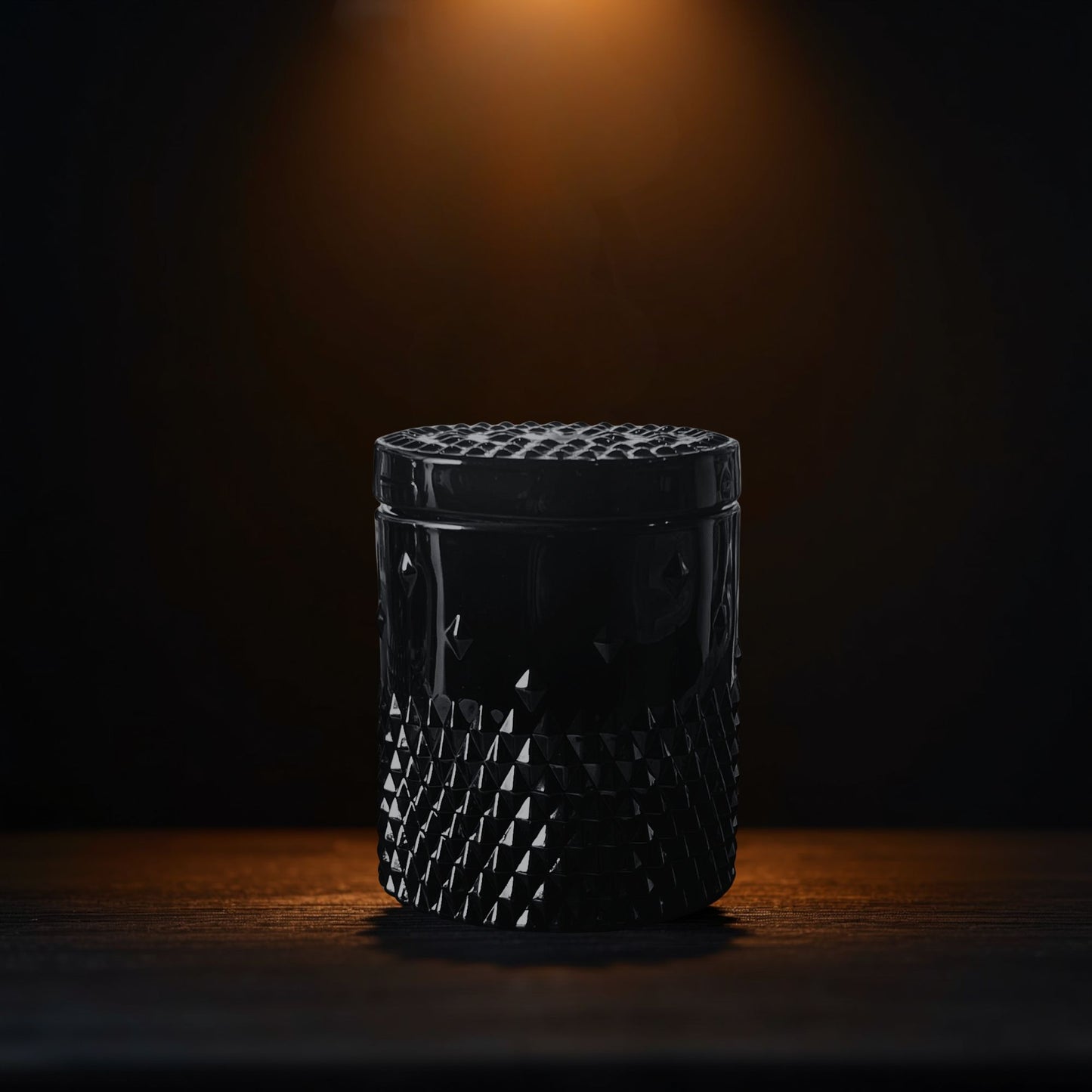 A black rockstud candle with a glossy faceted texture, lit by a warm spotlight on a dark wooden surface against a black background.