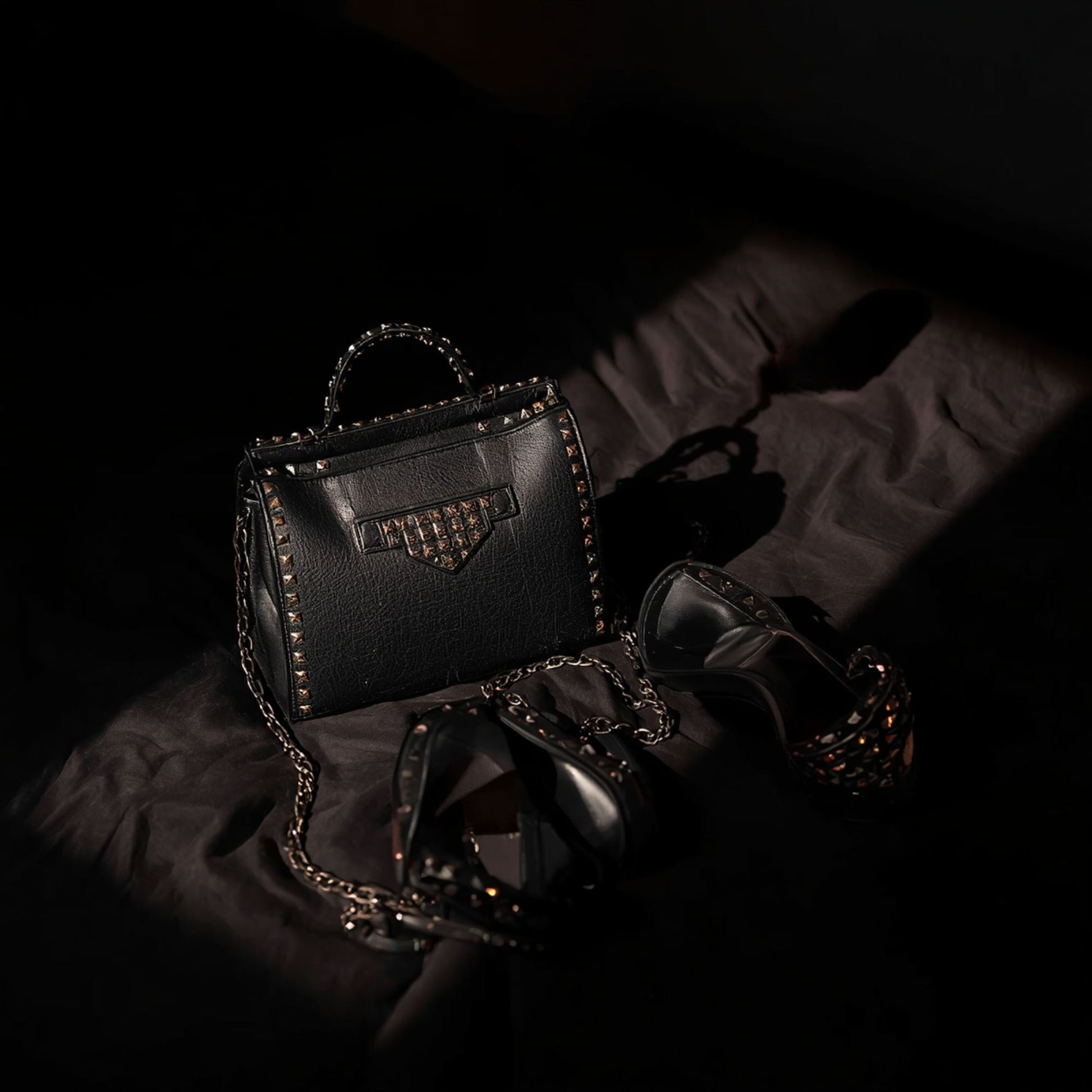 A black studded handbag and black high‑heeled shoes arranged on a dark fabric surface, lit by warm dramatic lighting that highlights the metallic studs and leather textures.