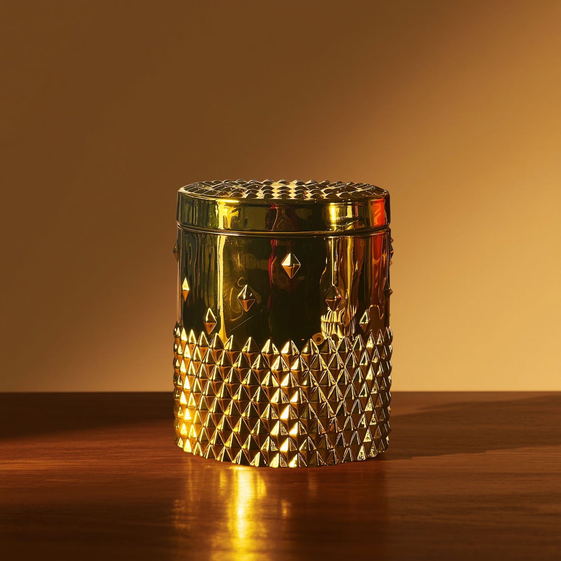 A gold rockstud candle with a reflective faceted texture, shown on a wooden surface with warm lighting highlighting the metallic finish.