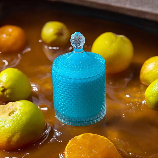 A deep green textured candle jar with a faceted lid, surrounded by sliced citrus in a glossy liquid, representing the Metaphor Man candle.