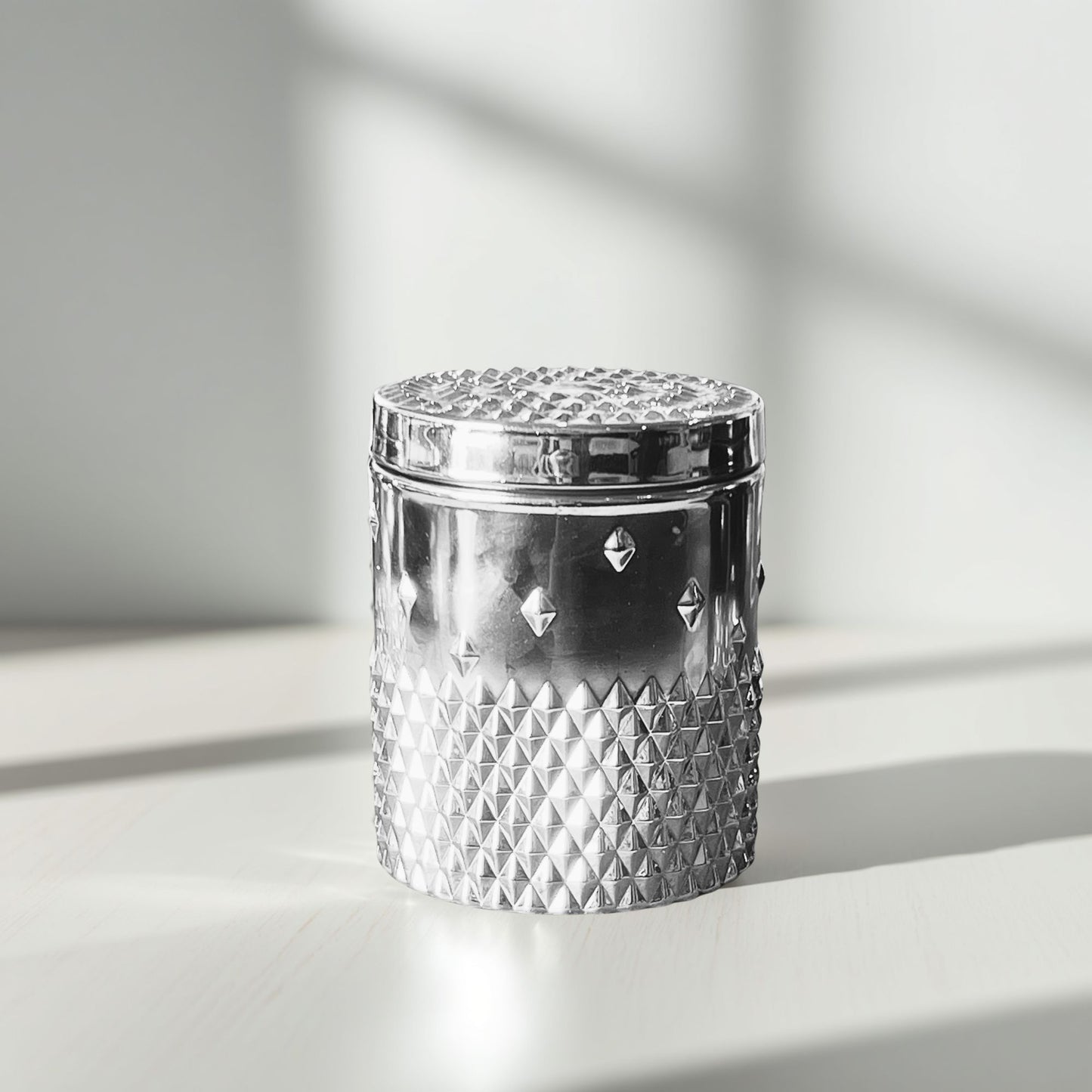 A silver rockstud candle with a reflective diamond‑pattern texture, shown on a light surface with bright side lighting highlighting the metallic finish.
