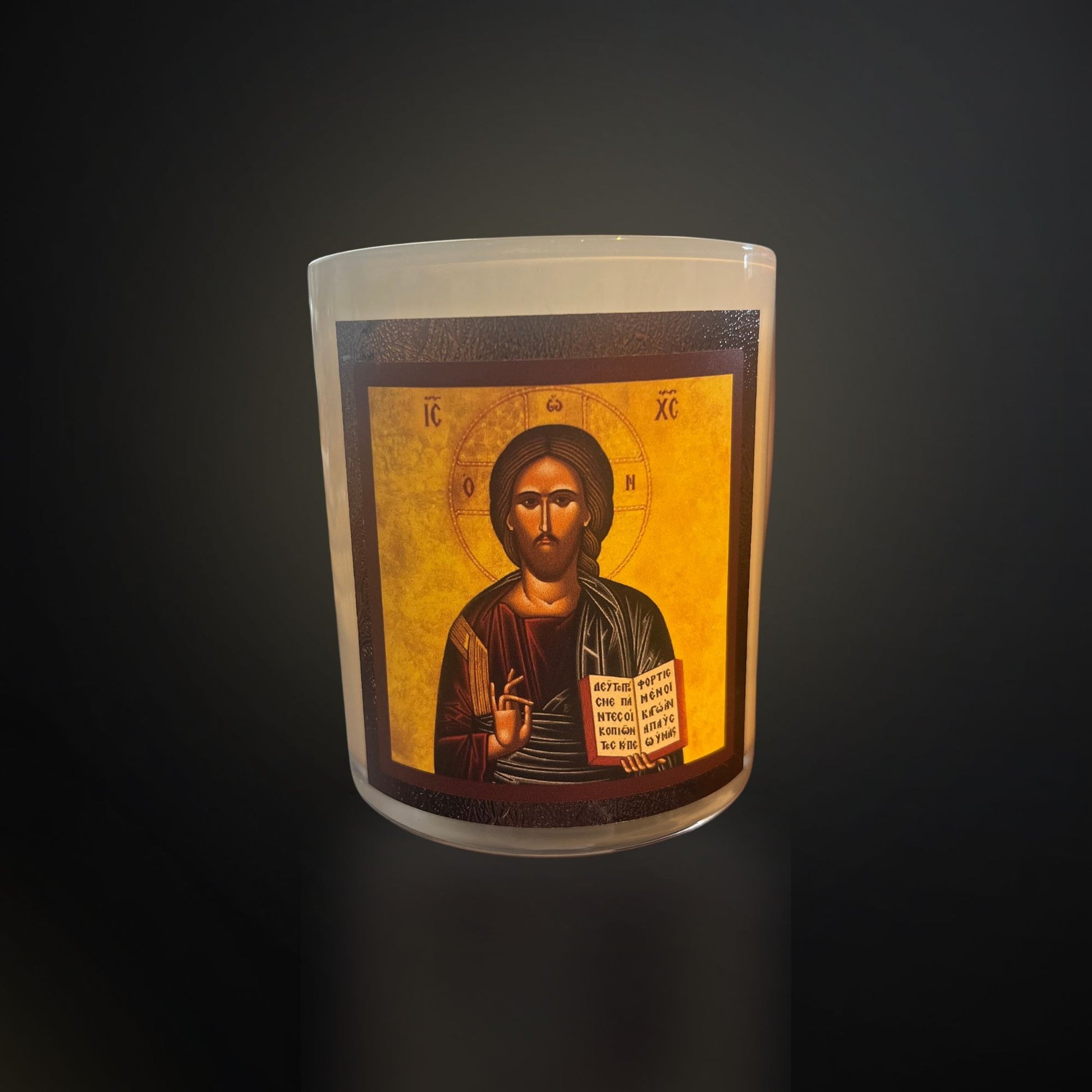 A white devotional candle featuring a detailed Christ icon, showing Jesus with a halo, blessing gesture, and an open book, photographed against a dark background.