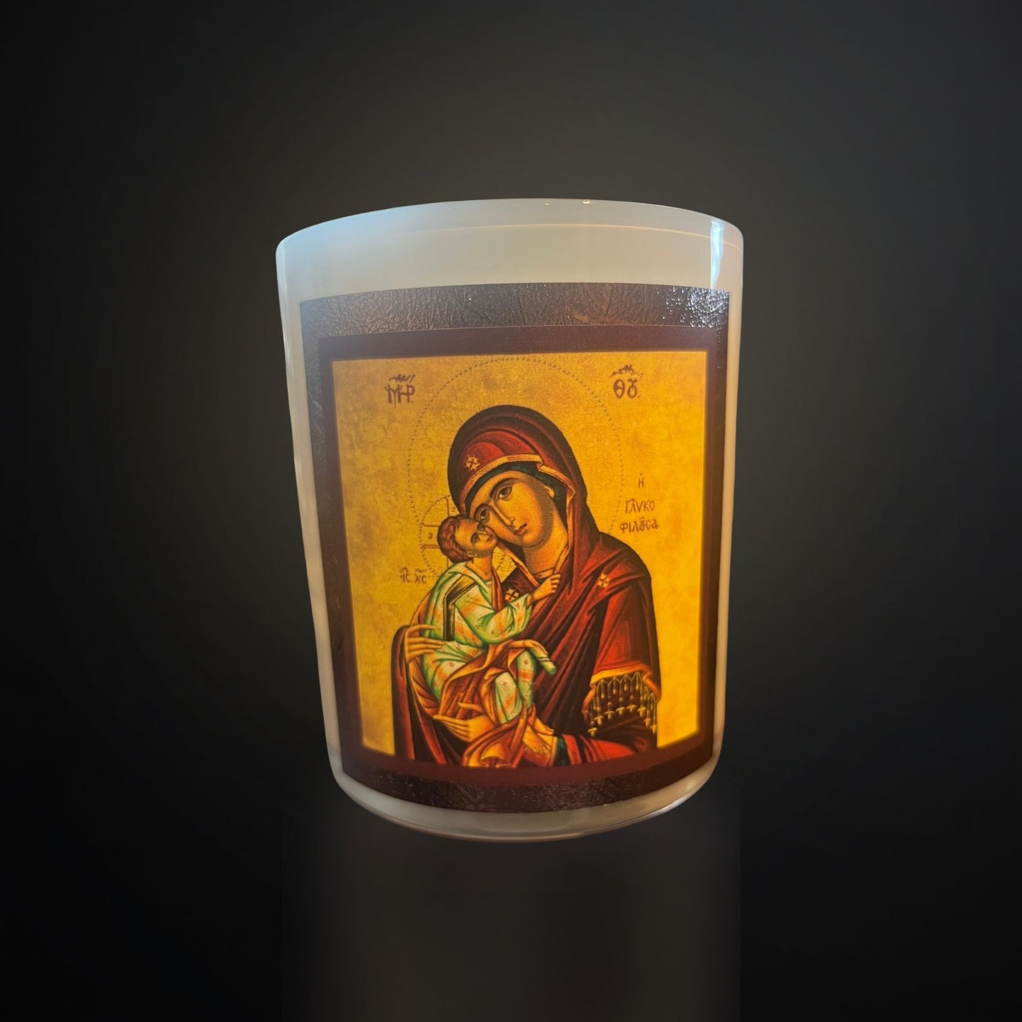 A white devotional candle featuring a detailed Mary icon holding the infant Jesus, both with halos, framed in warm tones against a dark background.