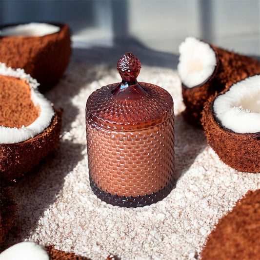 A textured glass candle jar resting on a bed of shredded coconut, surrounded by halved coconuts, representing the Brown Sugar & Coconut candle.