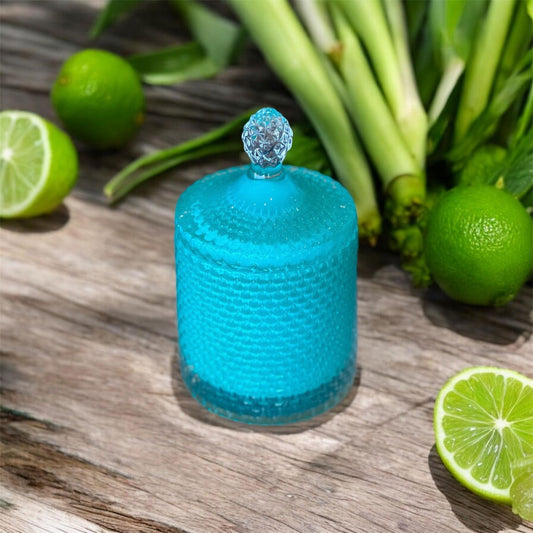 A deep teal‑green textured candle jar with a decorative lid, surrounded by fresh limes and lemongrass on a rustic wooden surface, representing the Lemongrass & Lime candle.