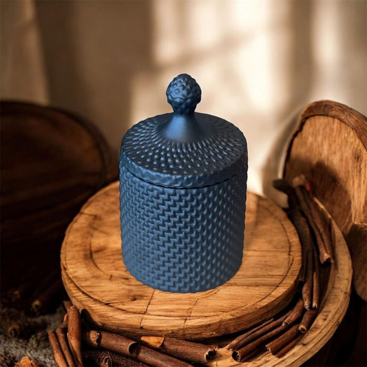 A dark blue textured candle jar with a decorative lid, surrounded by cinnamon sticks on a wooden surface, representing the Tobacco Vanille candle.