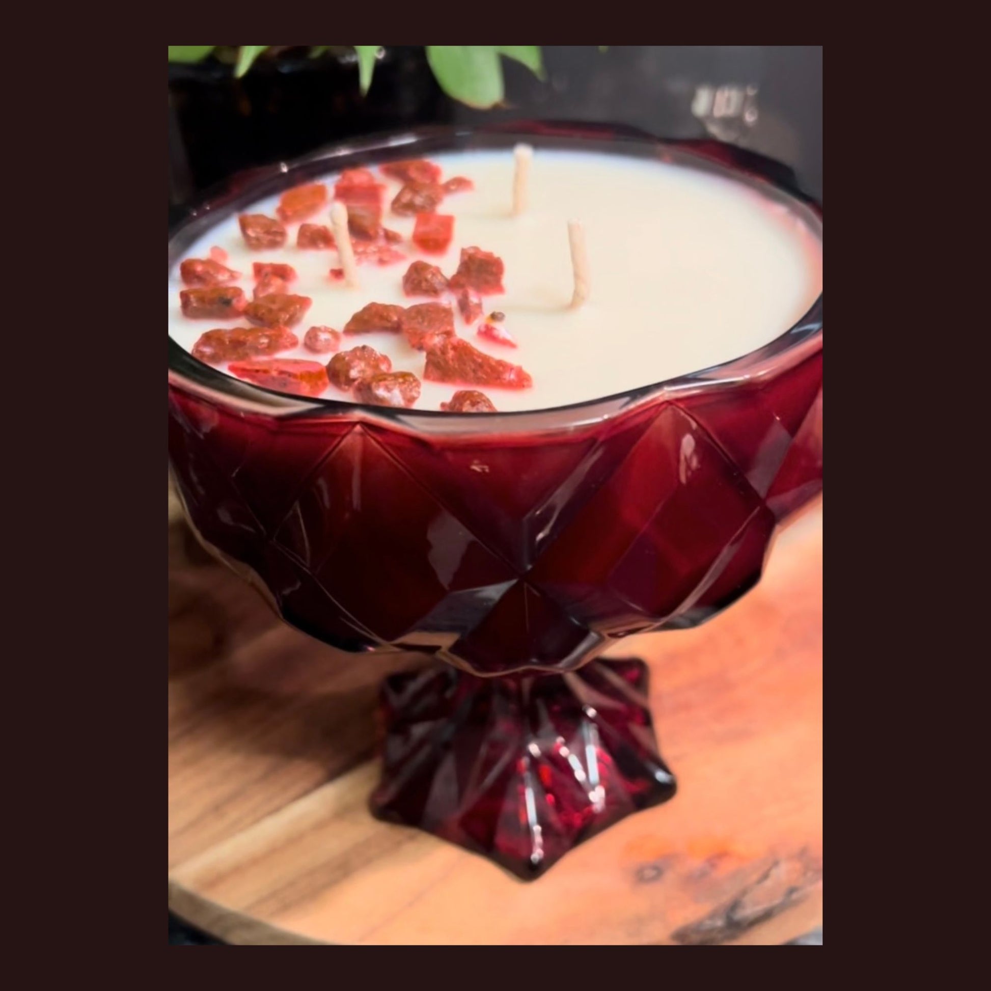 A ruby red chalice candle with white wax and red rose Livani pieces, placed on a wooden board with greenery behind it.