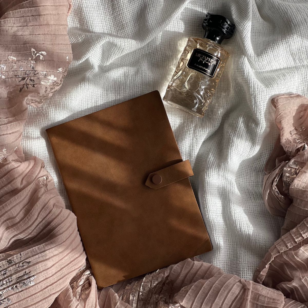 “Metaphor’s rose gold extrait bottle resting on embroidered silk and textured linen, surrounded by soft shadows and a leather-bound journal — evoking scent as memory.”