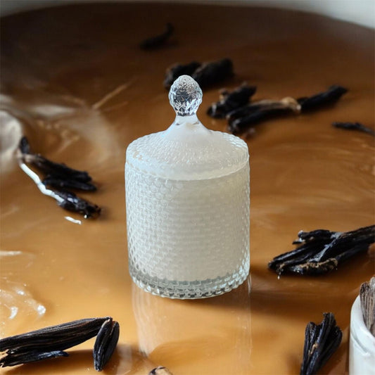 A textured glass candle jar resting on a glossy caramel surface, surrounded by dried vanilla beans, representing the Caramel Vanille candle.